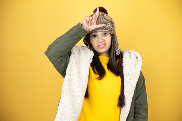 Young beautiful woman wearing a hat and a green winter coat over yellow background making fun of...