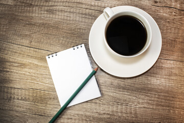 On a wooden texture table is a notepad with space for text near a white cup of coffee. Concept photo