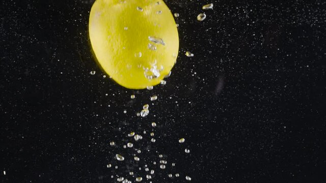 Close-up of whole lemon falling into water. Action. Whole lemon falls into water with bubbles on black background. Refreshing lemon drops into water