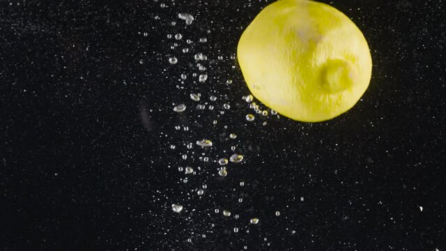 Close-up of whole lemon falling into water. Action. Whole lemon falls into water with bubbles on black background. Refreshing lemon drops into water