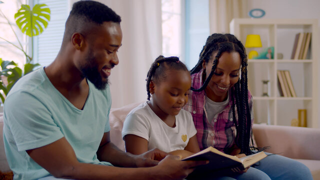 Cheerful african parents and little girl sitting on couch in living room reading book together