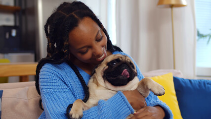 Smiling african woman playing with pet pug dog at home sitting on couch
