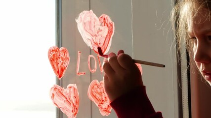 Little girl holds paintbrush in hand and drawing red heart on window glass. Valentine's day, love, dating, quarantine family leisure. Stay home art concept. Child painting playing indoors. New normal.