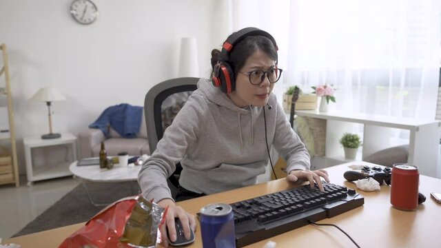 delighted young girl playing video games on computer wearing headphones. asian japanese female otaku wearing headsets and raising arms celebrating win of online fun. cheerful woman gamer lifestyle.