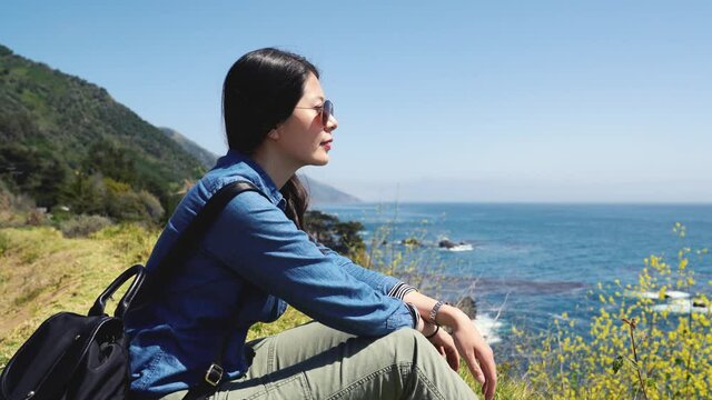 Young asian woman tourist wear sunglasses sitting on rock and looking at seashore. girl hiker carry backpacker relaxing on meadow in big sur route 1 trip california usa. lady love nature wild life.