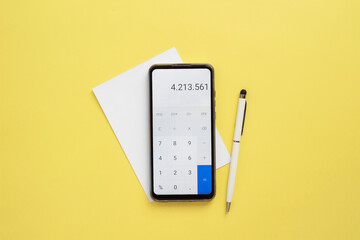 Calculator on phone, a pen and a blank white paper on yellow background.