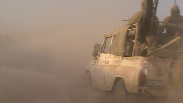 Islamist militants with machine guns and a black flag drive a car along a mountain road, the dust of the desert.