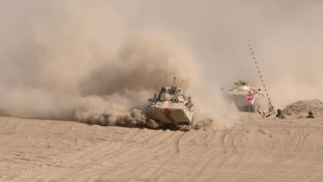 A column of armored vehicles passes through a checkpoint to perform a combat mission in a military conflict zone, dust rises from under the wheels.