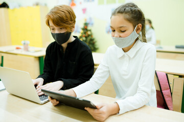 Students in medical masks sit at the table and take part in an online lesson using gadgets