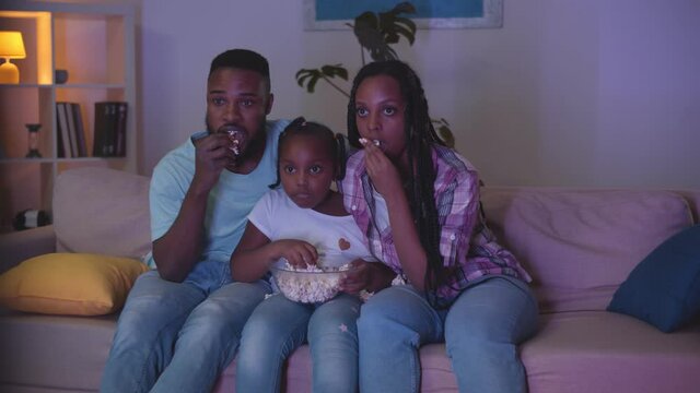 Happy african mom, dad and child sitting on sofa, eating popcorn and watching tv