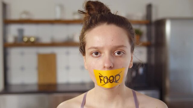 Young woman with stick tape with crossed out word food covering mouth standing at home