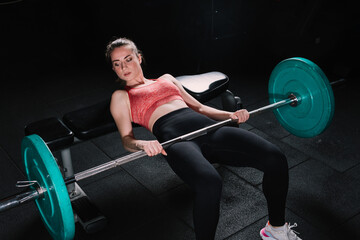 Young beautiful woman doing hip thrust. She poses with barbells in the gym.
