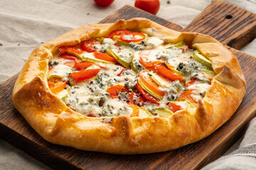 Homemade savory galette with vegetables, wheat pie with tomatoes, zucchini, blue cheese Gorgonzola. Rustic crust crostata on dark linen textile tablecloth.