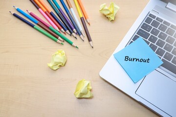 A piece of paper with a message on Burnout on the laptop keyboard with a group of color pencils and crumpled paper on the wooden table 