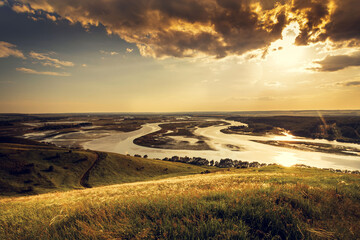 Fototapeta premium Sunny summer landscape with river, fields, green hills and beautiful clouds in blue sky. Pretty view of summer scenery on a nice day. Green trees growing along the river.