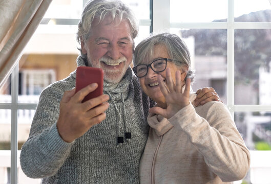 A beautiful couple of senior people hugging in front of the window on video call with distant family. Two active and smiling retirees