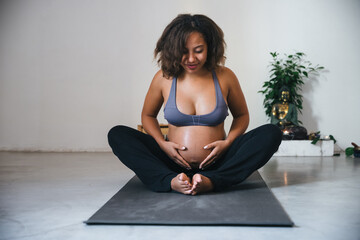 Young pregnant millennial woman sitting on the mat touches her belly after performing prenatal and meditation exercises at a yoga class - Concept of life and maternity - 402339427