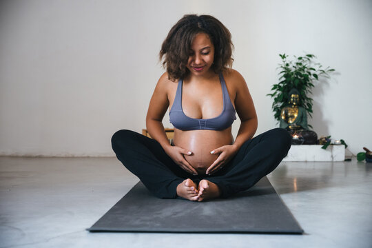 Young pregnant millennial woman sitting on the mat touches her belly after performing prenatal and meditation exercises at a yoga class - Concept of life and maternity