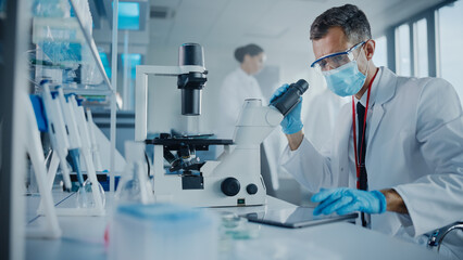 Medical Development Laboratory: Scientist Wearing Face Mask Looking Under Microscope, Enters Data...