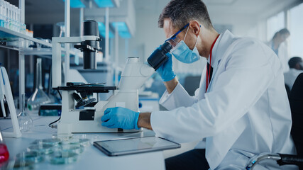 Medical Development Laboratory: Scientist Wearing Face Mask Looking Under Microscope and Using...