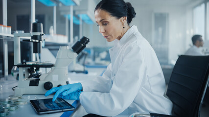 Medical Development Laboratory: Portrait of Beautiful Caucasian Female Scientist Using Microscope and Enters Data into Digital Tablet. Medicine, Biotechnology Research in Advanced Pharma Lab