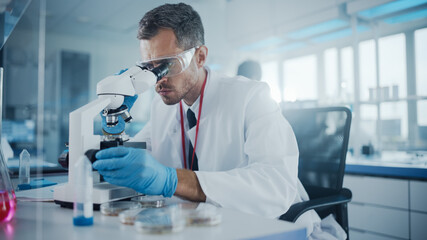 Medical Development Laboratory: Male Scientist Looking Under Microscope, Analyzes Petri Dish...