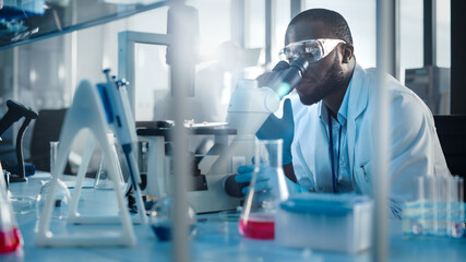 Medical Development Laboratory: Black Male Scientist Looking Under Microscope, Inspecting Petri Dish. Professionals Working in Advanced Scientific Lab doing Medicine, Vaccine, Biotechnology Research
