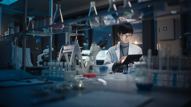 Medical Development Laboratory: East Asian Scientist Uses Digital Tablet Computer, Microscope, Conducting Experiment. Pharmaceutical Lab for Research Medicine, Biotechnology. Evening Work
