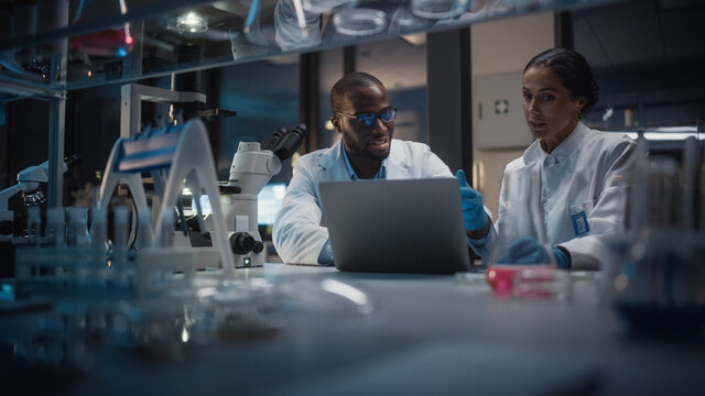 Modern Medical Research Laboratory: Caucasian and Black Scientists Work on Laptop, Do Data Analysis, Talk. Advanced Scientific Pharmaceutical Lab for Medicine, Biotechnology Development. Evening Time