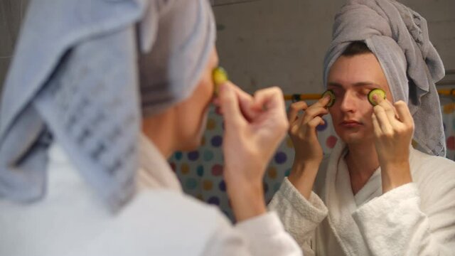 Young man in bathrobe and towel on head scrubbing face with cucumber slices in bathroom