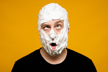 Studio portrait. A crazy man looks at the camera with his eyes rolled up, his face covered in shaving foam. On a yellow background