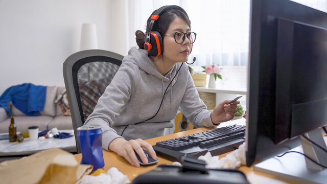 asian chinese young girl indoorsy holding credit card playing online gambling on desktop computer. lazy woman geek in headphones and glasses using debit card paying game cash. e commerce concept.