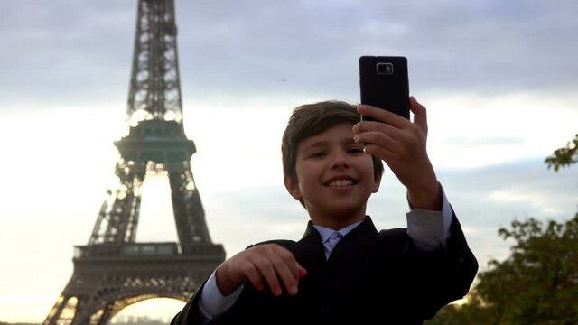 A smiling teenage boy in a black suit is taking a photo on the phone on the background of the Eiffel tower at the sunrise, Paris, France