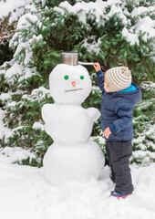 Happy child playing with a snowman outdoor in winter. Cute little child building a snowman and having fun in park or backyard.