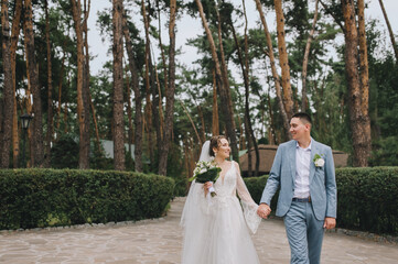 Fototapeta na wymiar Stylish groom in a blue suit and a beautiful bride in a white lace dress are walking holding hands in the forest, in nature with pine trees. Wedding portrait of the newlyweds.