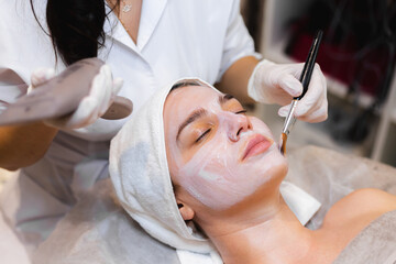 Beautician with a brush applies a white moisturizing mask to the face of a young girl client in a...