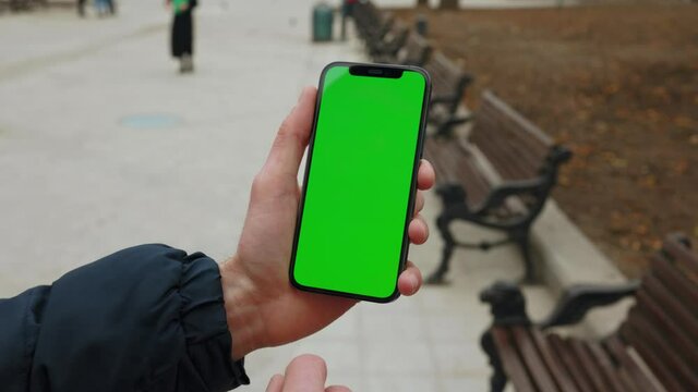 Young hands man holding use phone with vertical green screen in park background bench. Internet finger gadget device. Close up. Slow motion