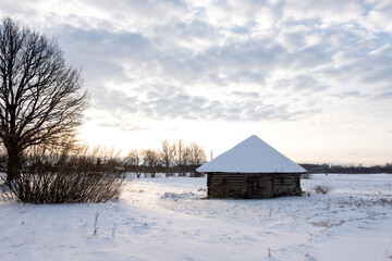 at sunset stands a snow - covered white barn made of wood in the Latvian countryside