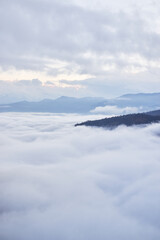Fototapeta na wymiar Low clouds, view of winter forest and mountains from observation deck, vertical picture of amazing natural phenomenon. Beautiful unreal panoramic view of mountains in clouds.