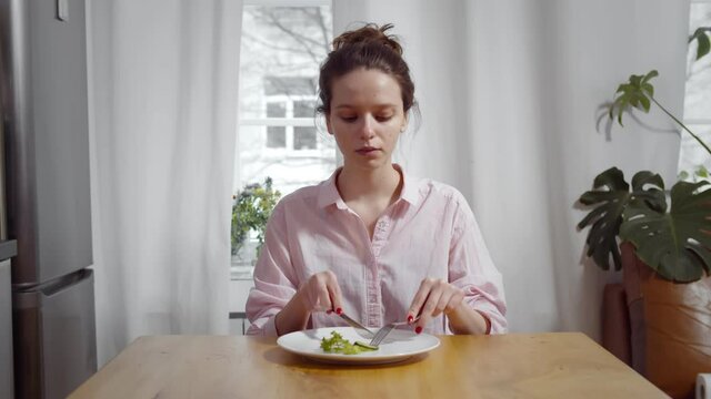 Young skinny woman sitting at table and eating piece of cucumber and salad with for and knife
