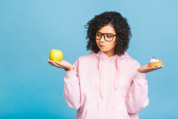 The girl makes the decision. American afro girl does not eat cake. Diet concept. Conception to lose weight. Isolated over blue background. Holding apple and cake.