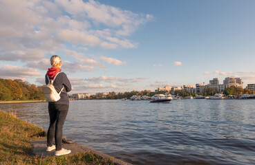 A young woman in casual comfortable clothes and with a backpack stands on the river bank admiring the view.