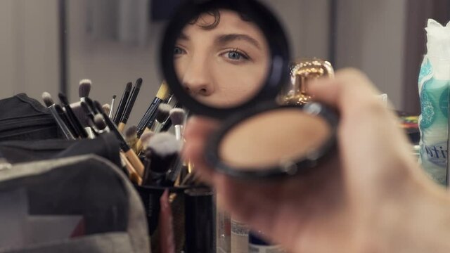 Young androgynous man with make-up looks into pocket mirror. Transgender man does a make up beauty routine. Close-up 4k