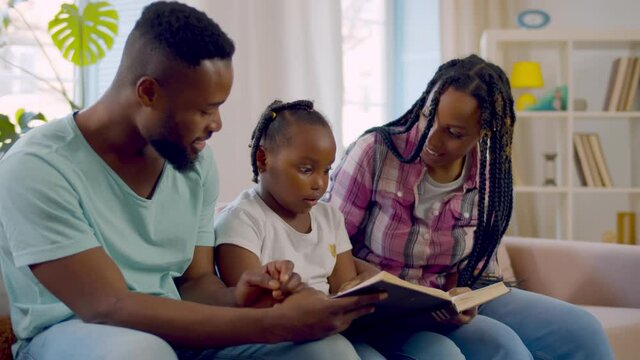 Cheerful african parents and little girl sitting on couch in living room reading book together