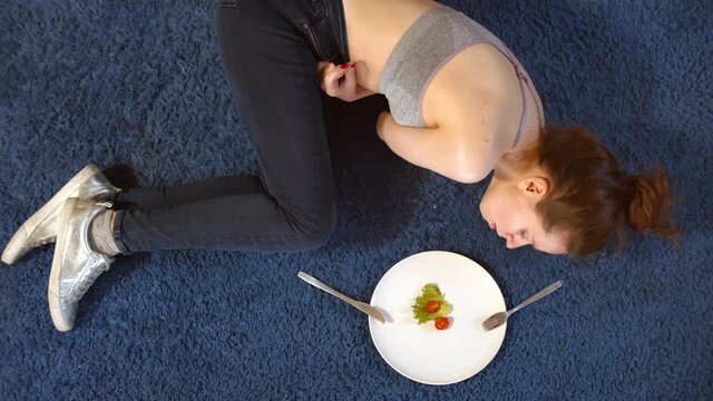 Sad woman having anorexia nervosa having small green vegetable and tomato on plate lying on floor