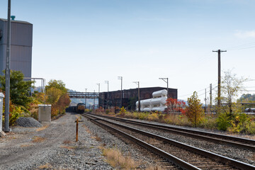 Train coming down a track running alongside industrial buildings in a rural area, fall season with autumn leaves, horizontal aspect