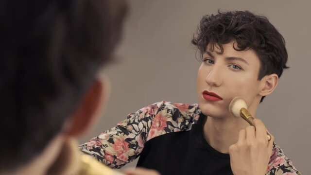 Young transgender man makes blush on the face using makeup brush. Androgynous guy does a make up beauty routine standing in front of mirror 4k