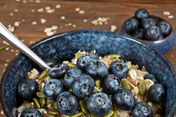 Oatmeal porridge with blueberries and seeds in a blue ceramic bowl with spoon on a wooden background.