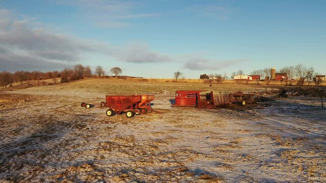 Establishing shot of Midwestern farm in spring time. Wagons, equipment, agricultural field covered in snow in the foreground, red barns in the background. Aerial view of American countryside landscape