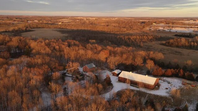 Establishing shot of Midwestern countryside in spring winter season. Aerial view of residential house, outbuildings in the forest. Sunny, soft sunlight at sunset